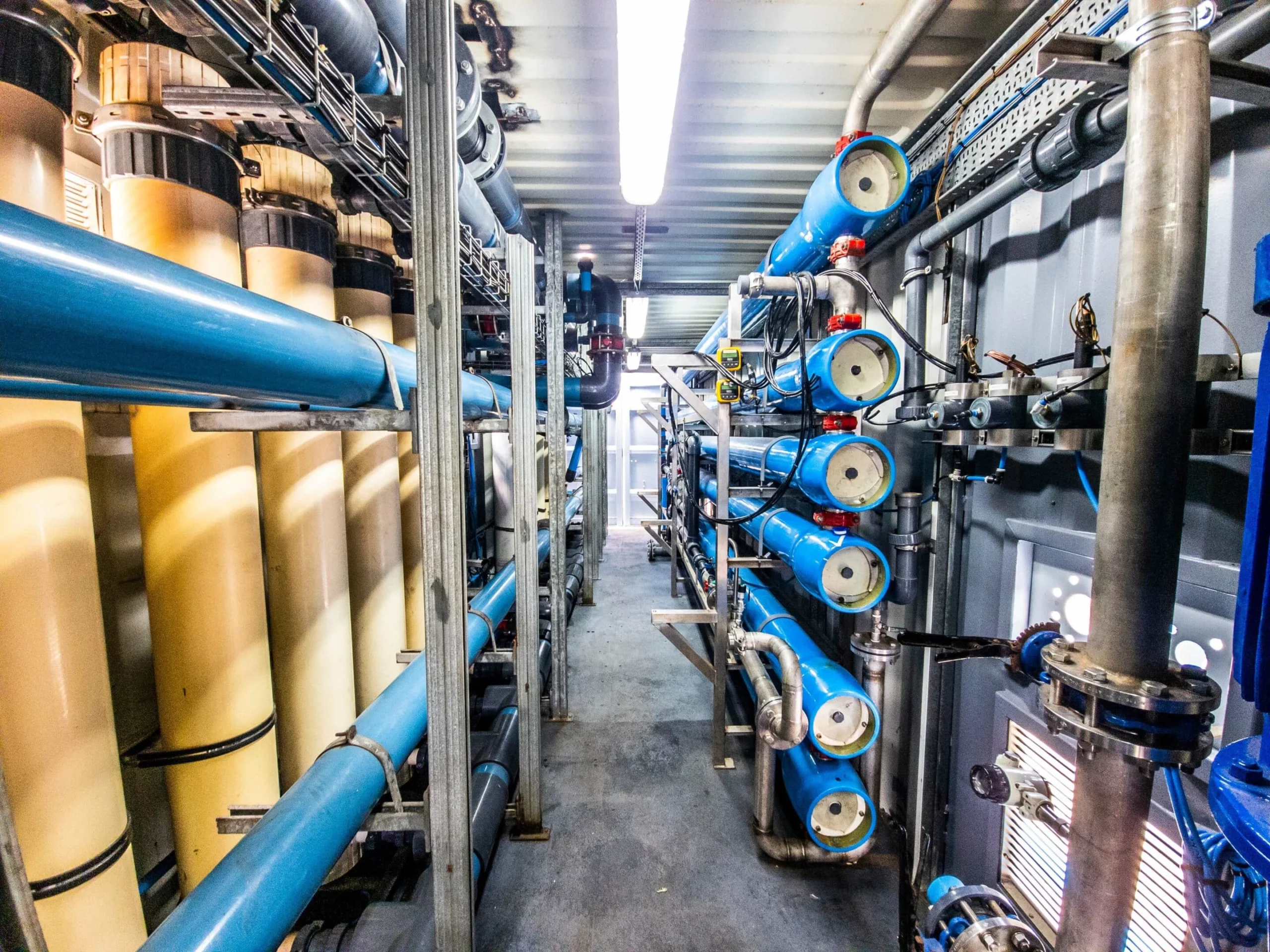 Interior View Of A Water Treatment System With Blue Pipes And White Cylindrical Units, Likely Reverse Osmosis Membranes.