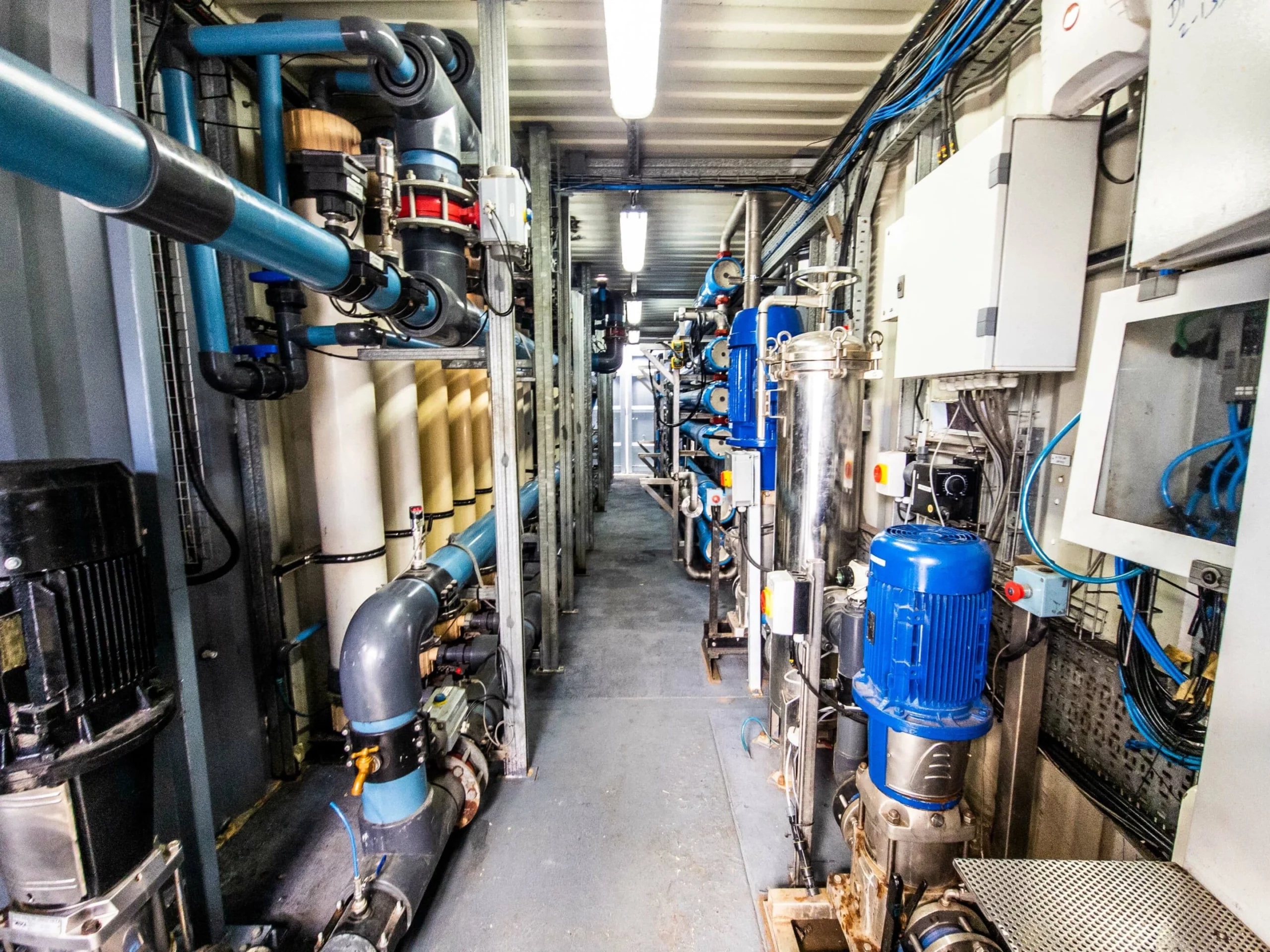 A Narrow Walkway Inside A Mobile Water Treatment Trailer Filled With Various Pumps, Pipes, And Control Equipment.