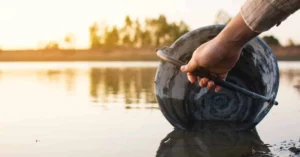 A Hand Scooping Water From A Murky Body Of Water With A Bucket, Symbolizing Water Scarcity.