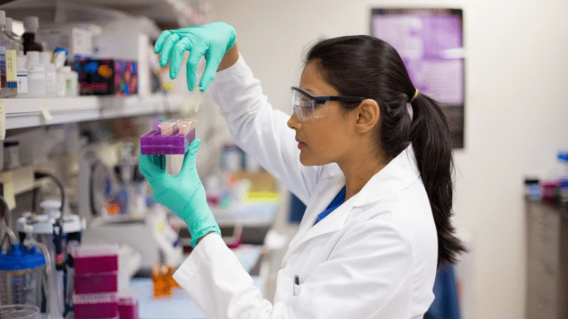 A Scientist In A Lab Coat And Safety Goggles, Wearing Teal Gloves, Carefully Examining Samples In A Laboratory Setting, Symbolizing Quality Control In Water Purification.